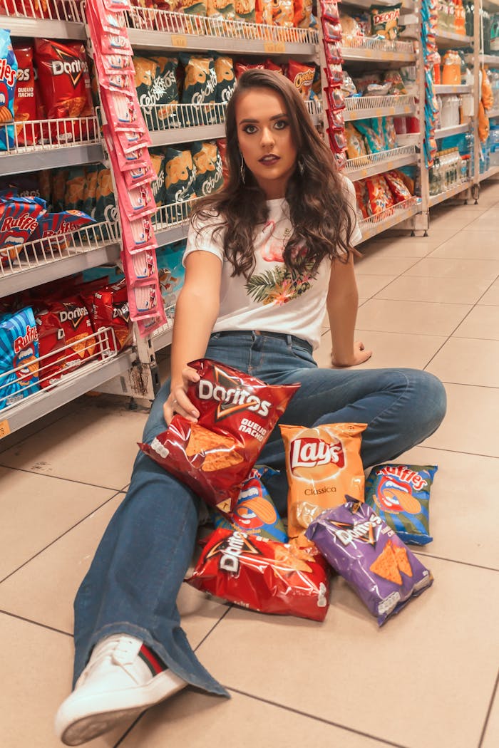 Young woman with trendy style sits on supermarket floor surrounded by snack bags.